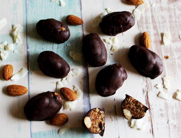 This photo is an overhead shot of healthy almond job bites dipped in chocolate. Almonds and coconut are sprinkled around them. One bite is cut in half to show the inside.