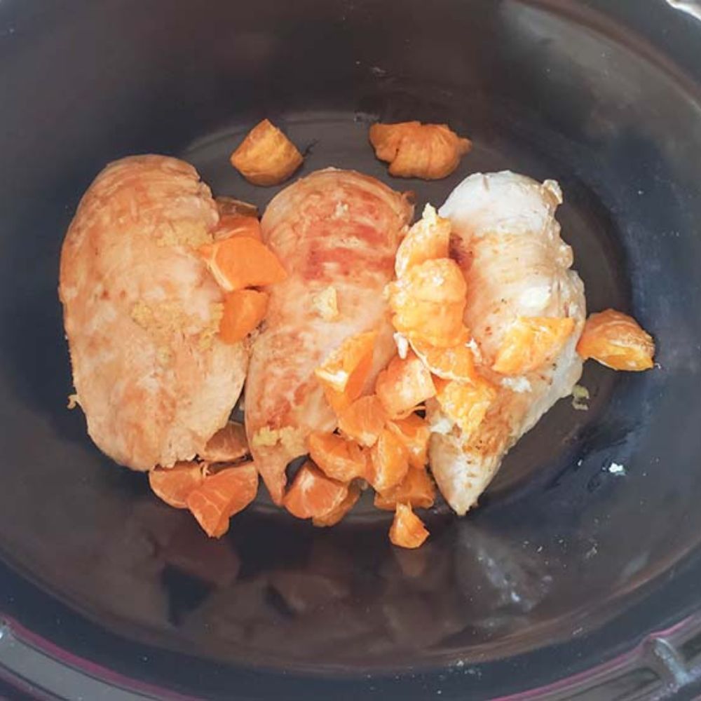 three boneless chicken breasts in a slow cooker with orange pieces on top