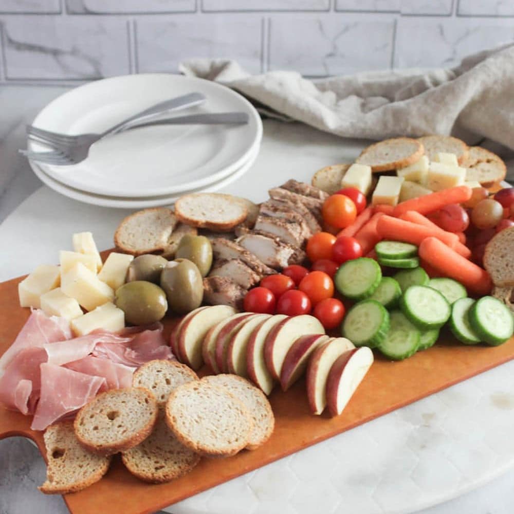 a picture of a fully completed charcuterie board filled with apples, vegetables, meats olives with two plates and forks on the side