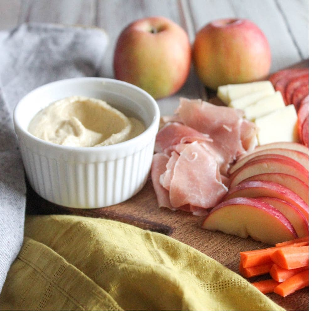 a close up of some of the ingredients on a charcuterie board: sliced apples, ham, hummus and cheese slices