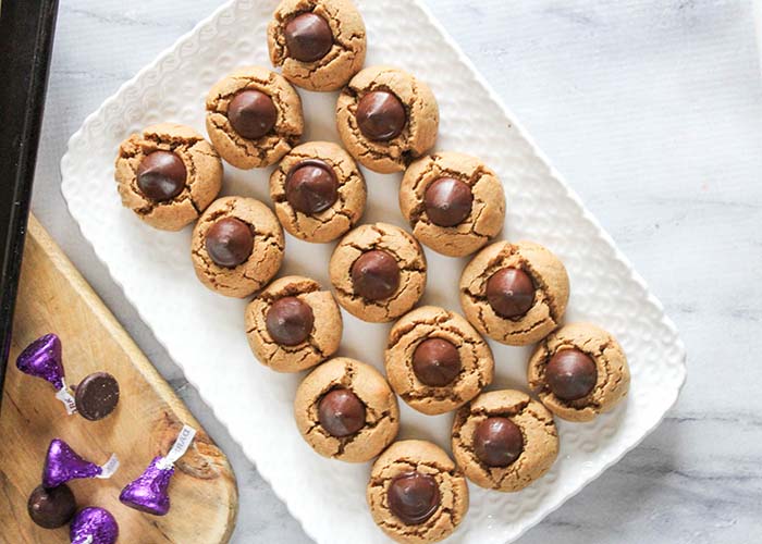 Overhead image of baked peanut butter blossom cookies with Hersey's kisses off to side. Cookies on white serving tray.