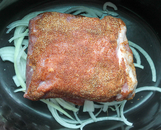 pork shoulder with spice rub on top of sliced onions in slow cooker