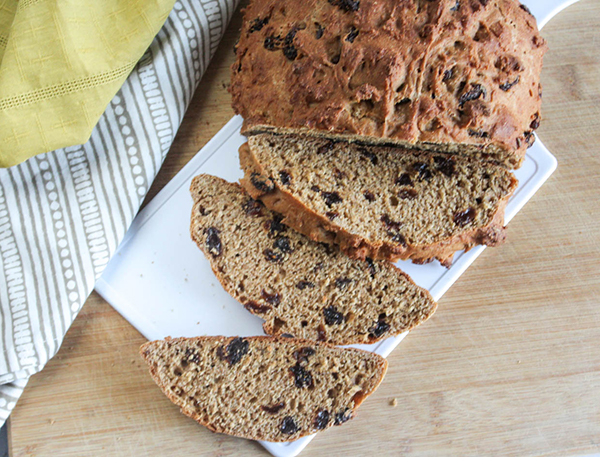 This picture is showing the finished cooked recipe of whole wheat Irish soda bread. The bread is sliced on a white cutting board with two dish towels, gray striped and yellow. 