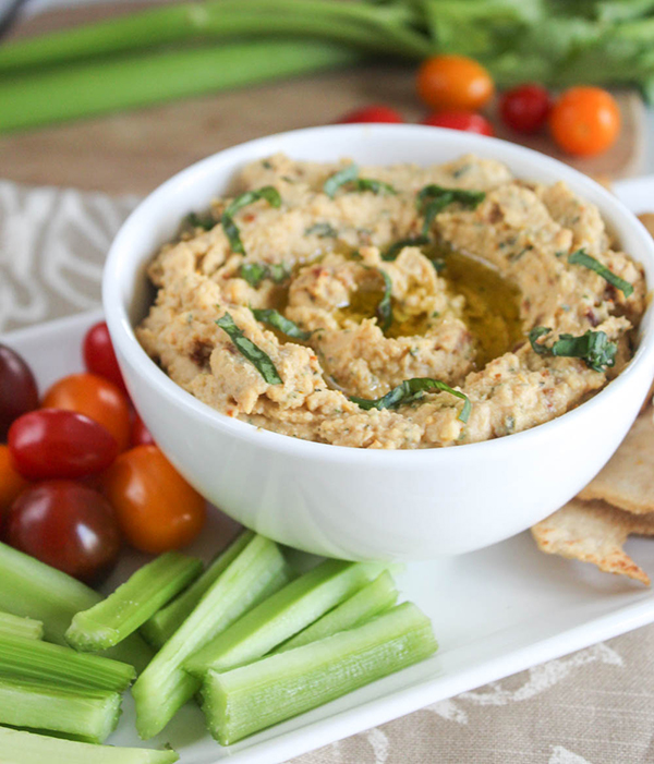 Hummus in serving bowl on white appetizer plate with celery, cherry tomatoes and pita chips.