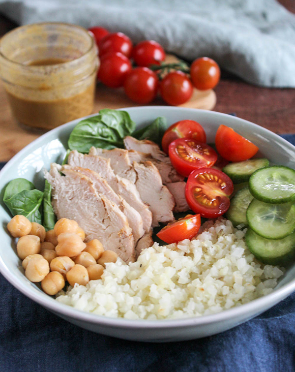 A bowl filled with grilled chicken, cauliflower rice, cucumbers and tomaotes and chickpeas