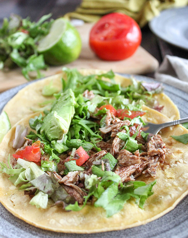 This image is a close up of finished chicken tostadas. Herbs, lime and tomato in the background of the picture on cutting board.