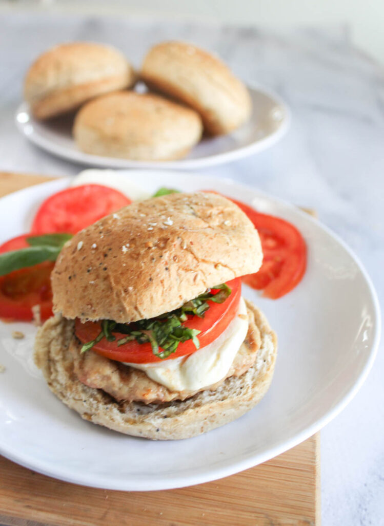 A burger on a roll with melted mozzarella cheese, basil and tomatoes with a plate of rolls in the background