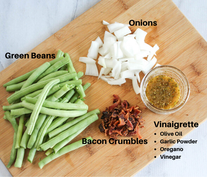 Ingredients to make green beans with onion and bacon on wooden cutting board, showing onions, green beans and bacon crumbles. Vinaigrette in bowl on side made of olive oil, garlic powder, oregano and vinegar.