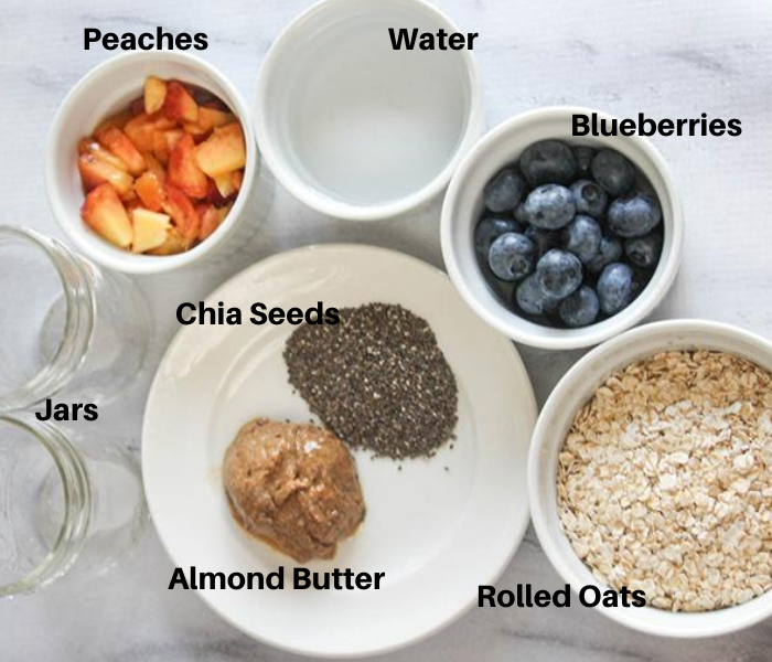 An overhead view of the ingredients in white bowls:  chopped peaches, water, blueberries, oatmeal, chia seeds, almond butter and 2 jars