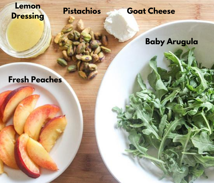 The ingredients for the salad, peaches, lemon dressing in a jar, pistachios, goat cheese baby arugula