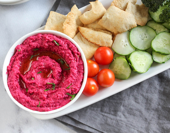 an overhead view of roated beet hummus in a bowl with fresh vegetables (tomatoes, cucumbers) and pita chips