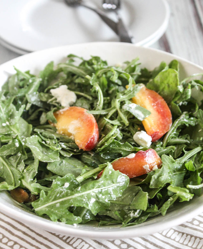 Arugula and sliced peaches in a white bowl with goat cheese and pistachios