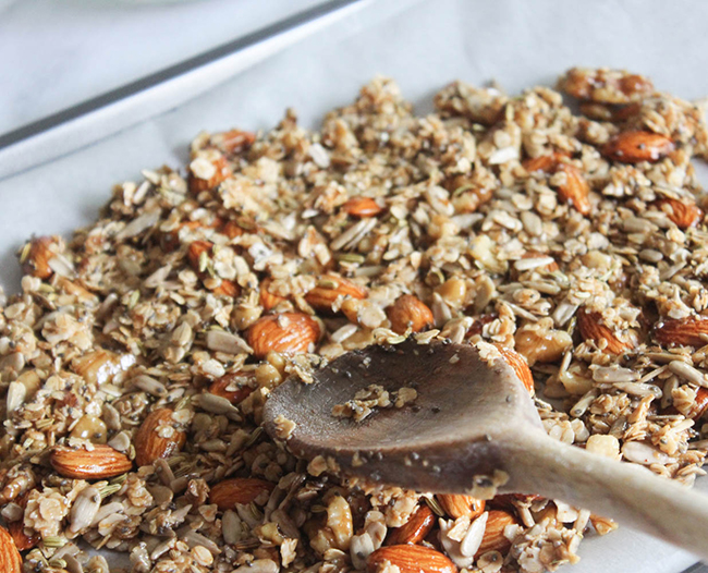 A picture of granola on a baking sheet with a wooden spoon pressing it down.