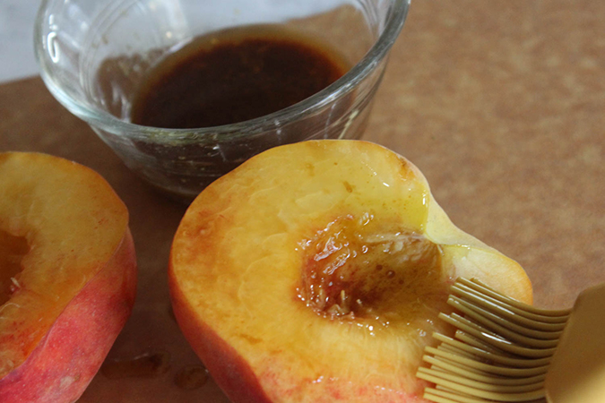 Step of recipe to demonstrate peaches being bruched with one tablespoon of marinade. Marinade is to the side in glass dish.