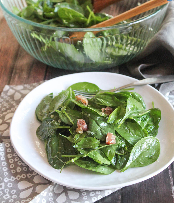 a close up picture of spinach salad on a plate with a glass salad bowl in the background with salad in it