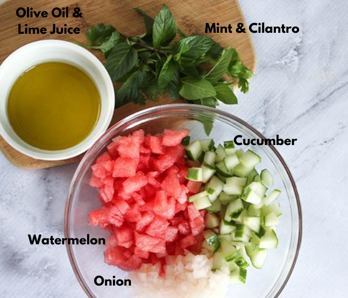 An overhead shot of all of the ingredients: olive oil & lime juice in a white bowl, fresh mint & cilantro on a cutting board and diced cucumbers, watermelon and onion in a glass bowl