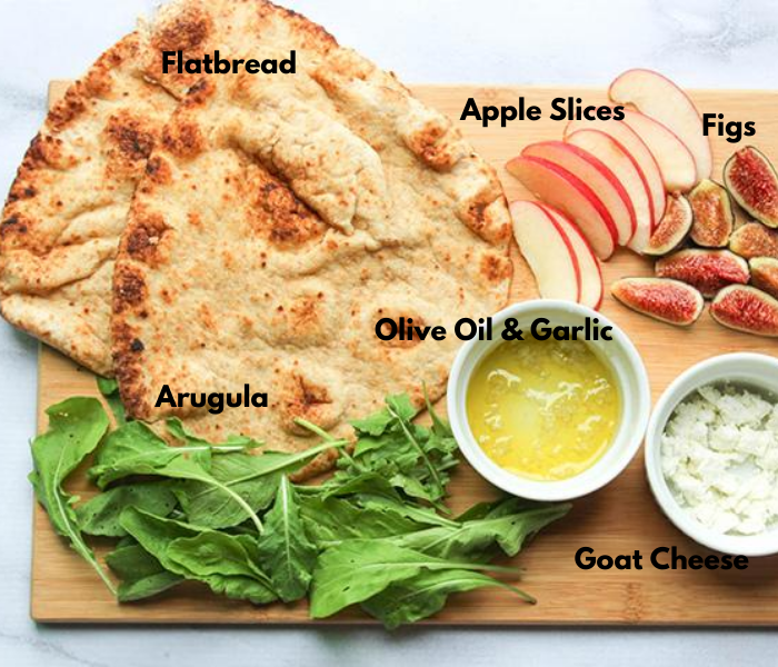 The ingredients on a cutting board: two naan breads, apple slices, fig slices, olive oil goat cheese and arugula