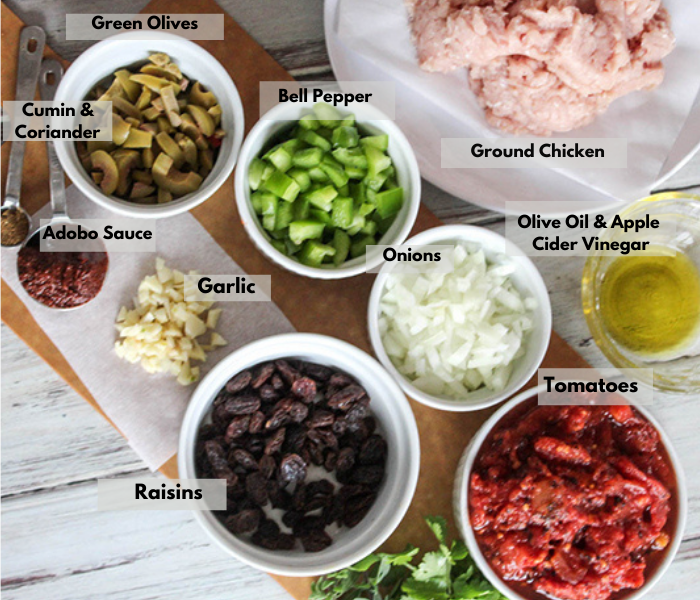 an overhead view of the ingredients: clockwise: bell peppers, ground chicken, olive oil, apple cider vinegar, tomatoes, cilantro, raisins, onions, garlic adobo sauce, cumin, corriander olives