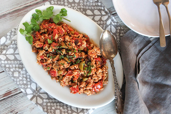 overhead view of the Chicken Picadillo in a white serving dish with serving spoon on side. Dish is placed on a table with natural brown linen napkin.