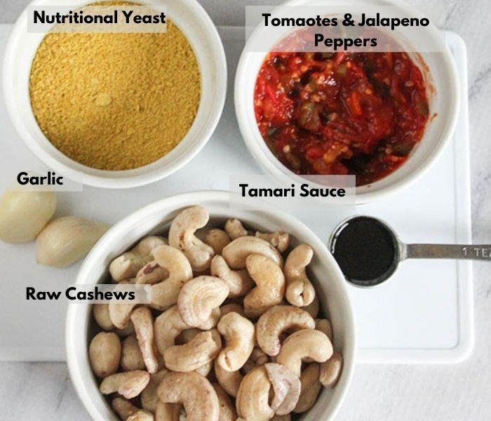 Ingredients for vegan queso dip. Garlic is on side. Tamari sauce is in teaspoon. In white bowls are tomatoes and jalapeno, nutritional yeast and raw cashews.