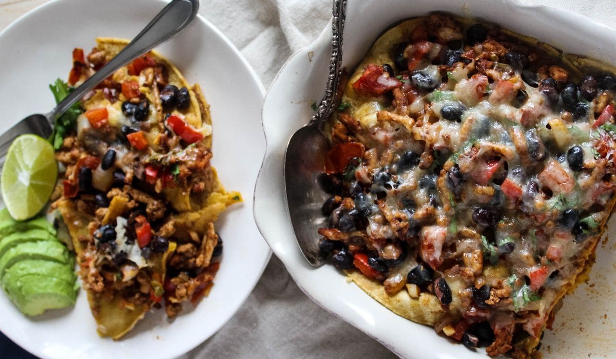 An overhead view of ground chicken casserole in a casserole dish with a spoon and a slice of the casserole next to it on a white plate with a fork, avocado slices and wedge of lime