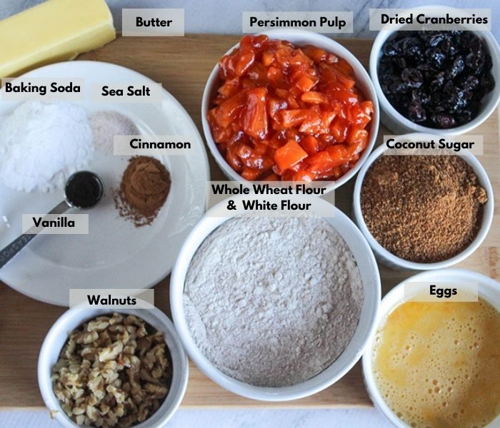 an overhead view of the ingredients: vanilla, cinnamon, baking soda sea salt, butter, persimmon pulp, dried cranberries, coconut sugar, eggs, flour, walnuts