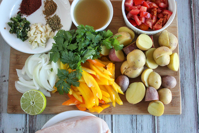 overhead view of Slow-Cooker Chipotle Chicken ingredients on wooden cutting board including spices, garlic, chicken broth, potatoes, cilantro, onions, peppers, lime