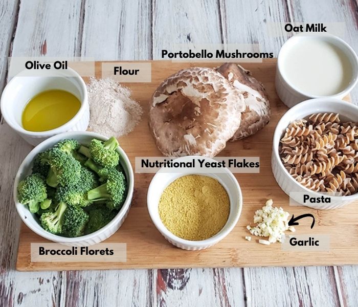 vegan mac and cheese ingredients on a cutting board: olive oil, flour, mshrooms, milk, broccoli, nutritional yeast, garlic and pasta