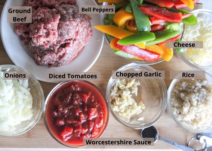 deconstructed peppers ingredients on a cutting board: tomaotes, onions, ground beef, bell peppers, rice garlic worcestershire sauce and grated cheese