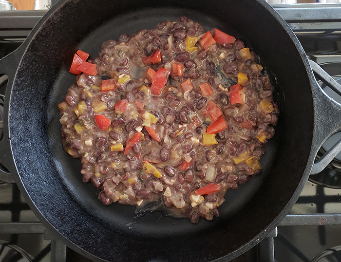 This image shows step four of the recipe. Black beans, water and cumin are added to the pan of cooked peppers and onions.