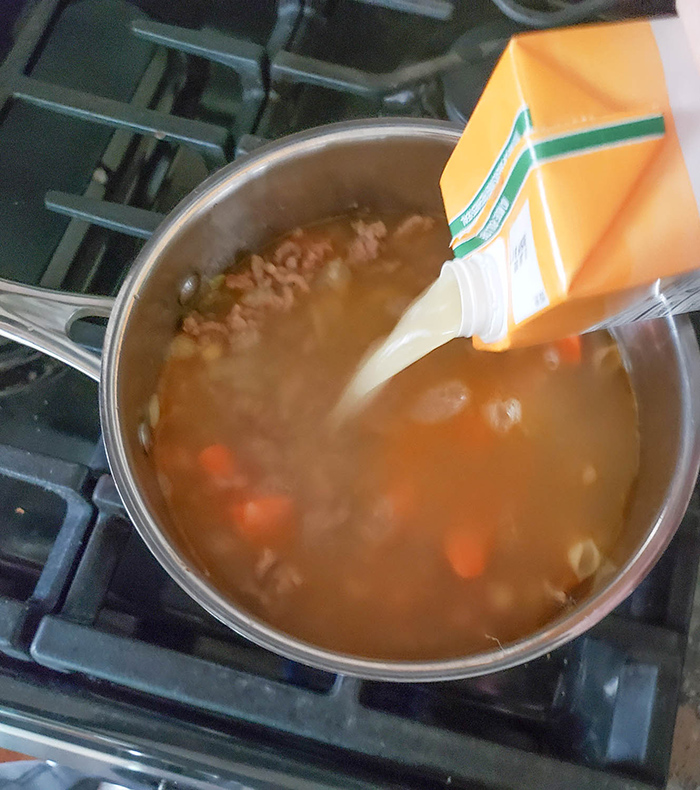 Chicken broth pouring into a pot of sausage and vegetables on a stove.