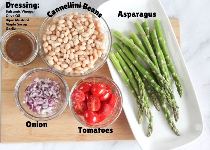 asparagus salad ingredients: asparagus, tomaotes, onion, dressing, white beans on a cutting board.