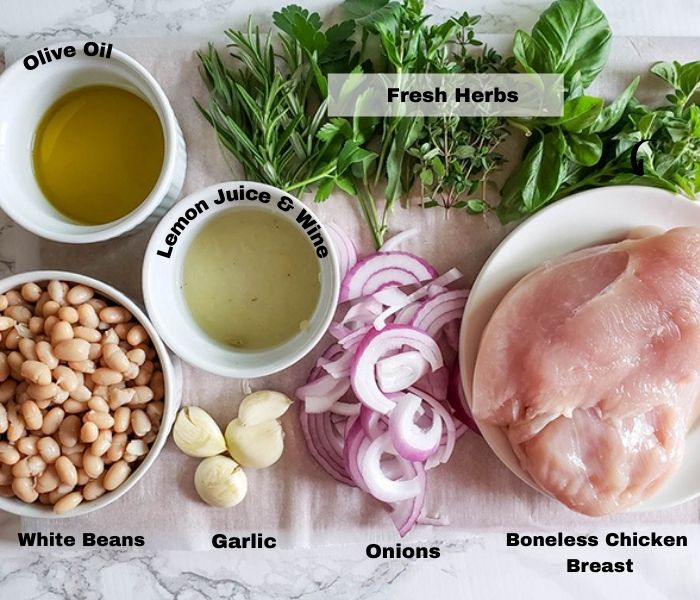 ingredients for Garlic Herb Chicken and Cannellini Beans on a cutting board with fresh herbs, olive oil, chicken, red onions, garlic, white beans and lemon juice and wine