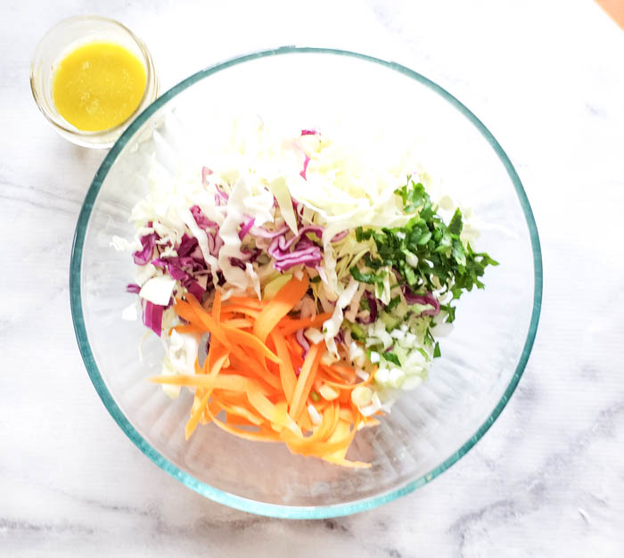 an overhead view of the coleslaw ingredients in a bowl, carrots, cabbage, green onions and herbs with the dressing in a jar on the side