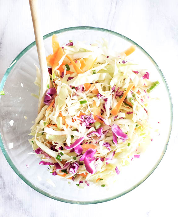 the coleslaw ingredients mixed together in a glass bowl with a wooden spoon
