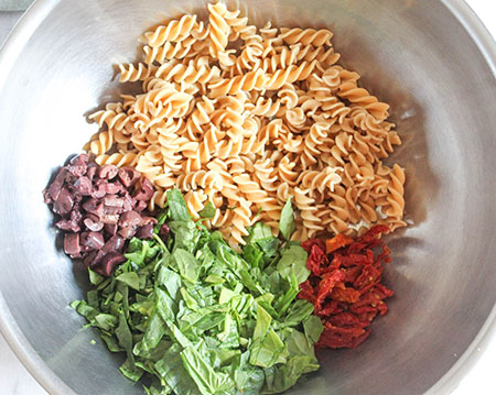 The pasta salad ingredients in a mixing bowl: chopped spinach, olives, sun-dried tomatoes and pasta