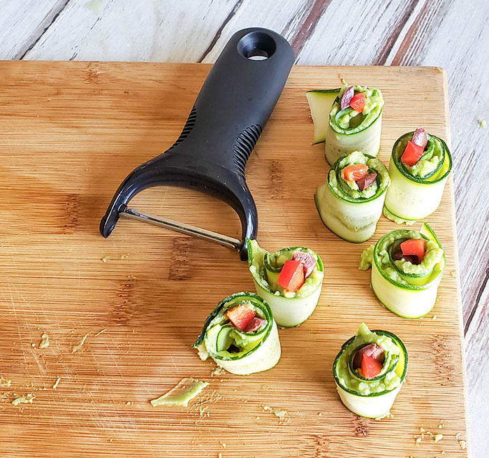 This image shows the final step of recipe. Zucchini rolls are topped with pieces of tomatoes. Seven zucchini rolls are ready to be plated and sitting on wooden cutting board beside vegetable peeler.