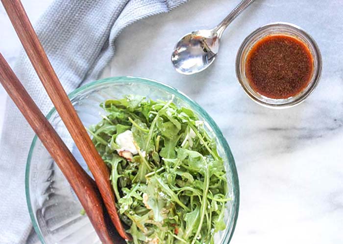 An overhead view of arugula salad with two wooden spoons in side and a side of dressing. This salad is ready to serve on a marble countertop.