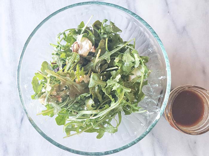 This photo shows how to mix the salad ingredients together and serve with a side of dressing. The arugula, pecans, figs and goat cheese crumbles are in a large glass salad bowl with a smaller glass bowl of dressing on the side.