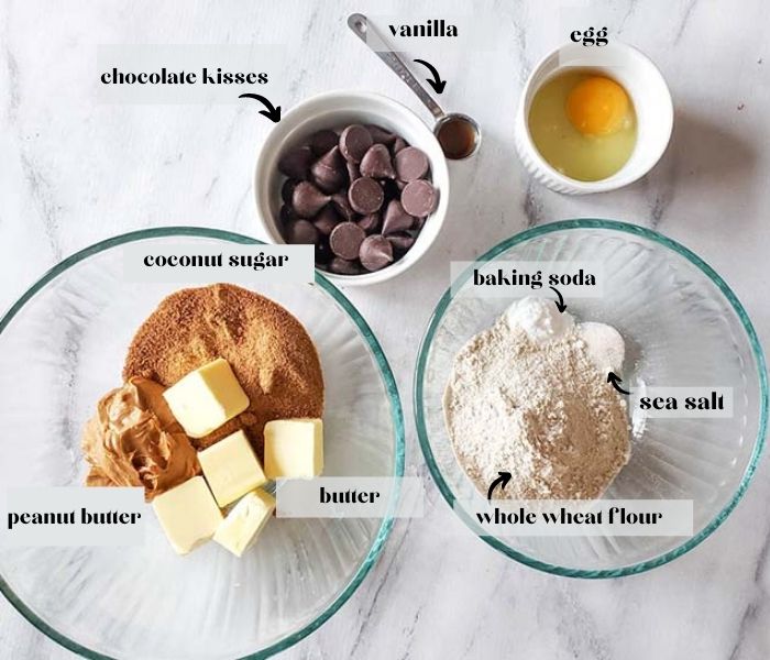 Overhead image of Peanut Butter Blossoms Cookies ingredients. Shown are coconut sugar, peanut butter and butter in a glass bowl. Baking soda, sea salt, whole wheat flour in second glass bowl. Chocolate kisses, egg and teaspoon of vanilla.