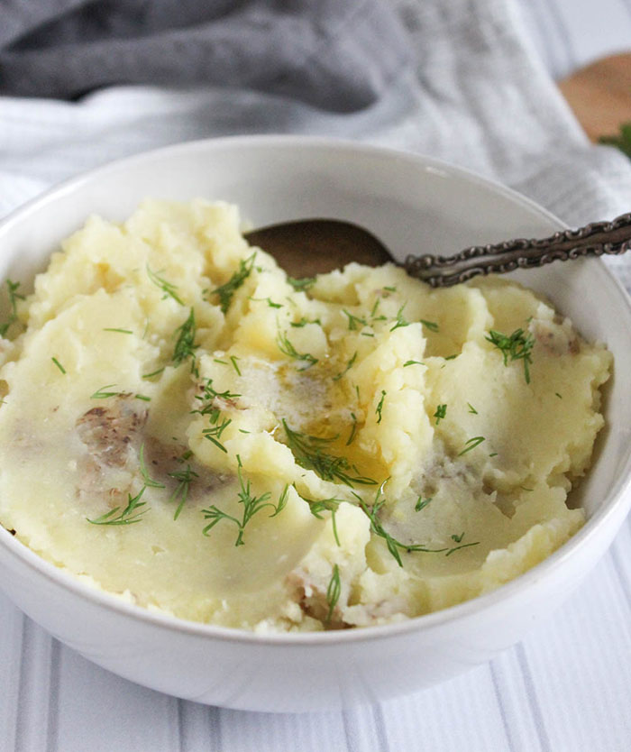 A close up picture of mashed potatoes in a white bowl with fresh dill and butter