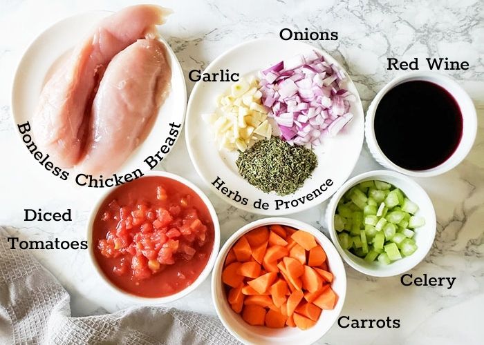 Ingredients for instant pot red wine chicken on countertop in white bowls. Ingredients pictured: boneless chicken breast, garlic, herbs, red wine, diced red onion, tomatoes carrots and celery.