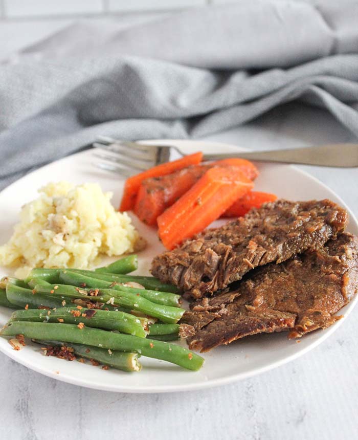 sliced pot roast on a white plate with green beans, mashed potatoes and carrots