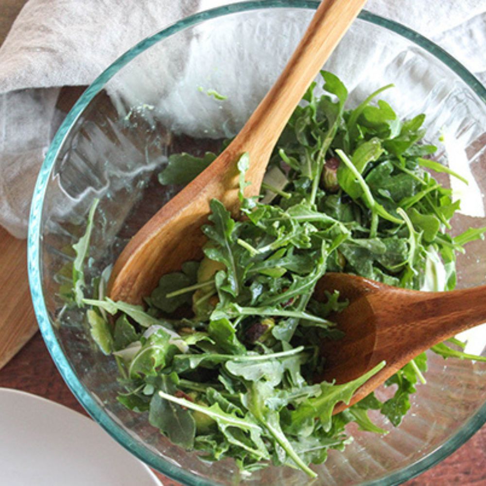 overhead view of arugula in clear glass mixing bowl wooden spoons tossing ingredients and dressing with linen towel to the side