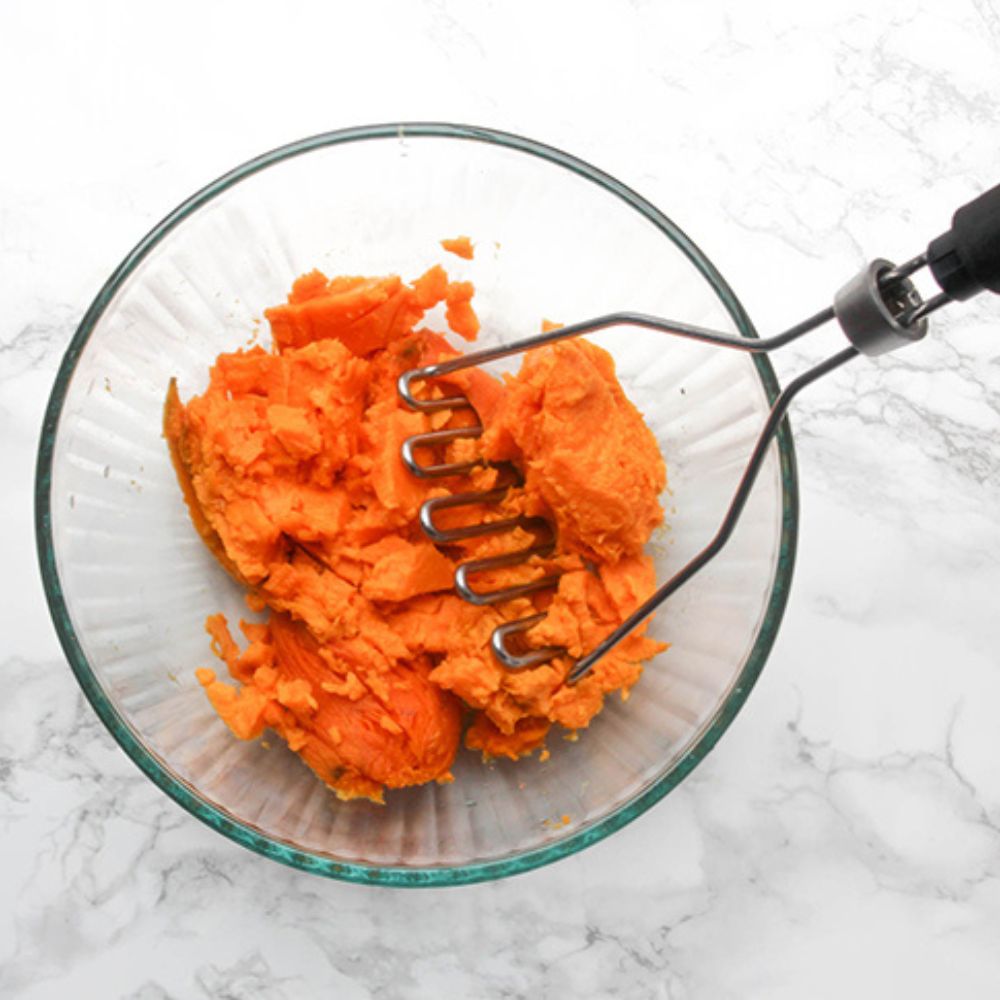 mashed sweet potatoes in a glass bowl with a potato masher