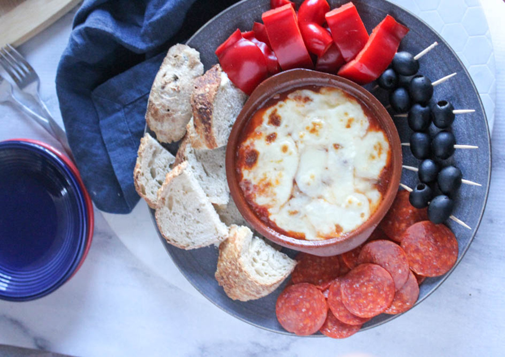 overhead view of pizza dip in a bowl with a plate of dippers bread, pepperoni, olives and peppers