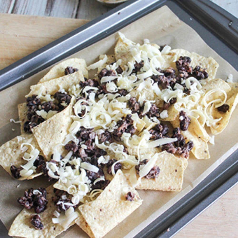 A picture of nacho chips on a parchment lined sheet pan topped iwth smashed black beans and shredded cheddar cheese.