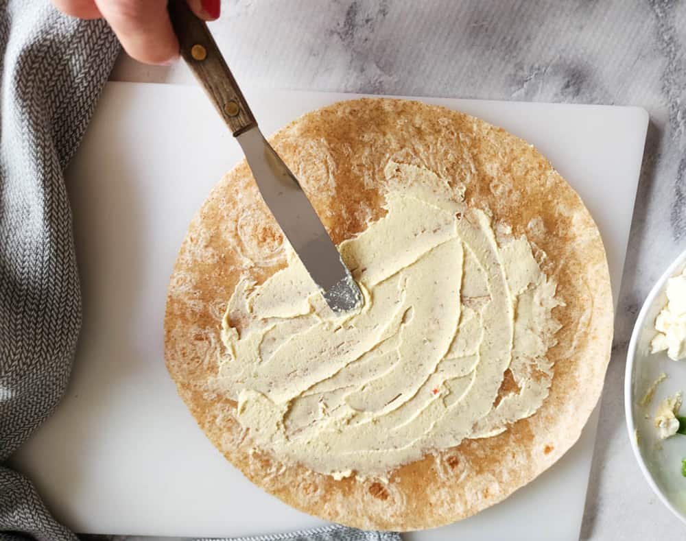 a process picture of a knife spreading hummus on a wrap