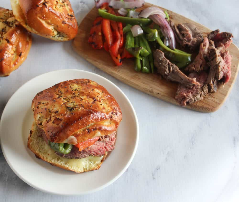 Overhead view of assembling Filet Mignon Sandwich with a sandwich on white plate and filet mignon, peppers and onions on wooden cutting board.