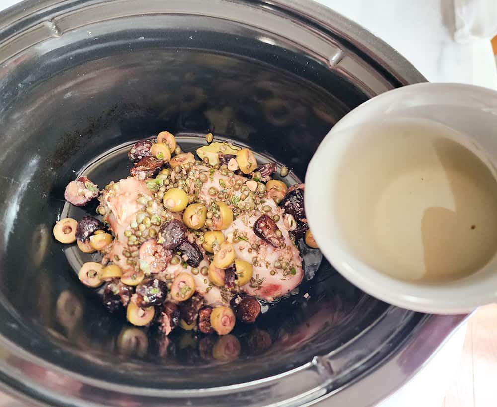 uncooked chicken marbella in a slow cooker with wine pouring in